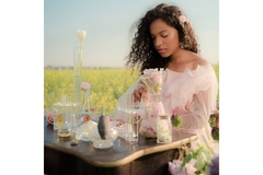 A woman mixes chemicals on a wooden desk decorated with flowers, sitting in a field of yellow canola flowers.